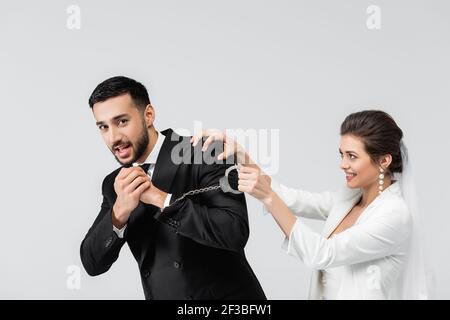 Lächelnde Braut hält Handschellen von verängstigten muslimischen Bräutigam isoliert auf Grau Stockfoto