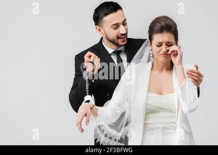 Lächelnder arabischer Bräutigam hält Handschellen in der Nähe weinende Braut isoliert auf Grau Stockfoto