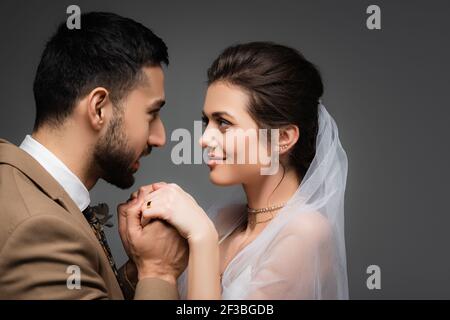 Eleganter muslimischer Mann, der die Hände einer glücklichen Braut isoliert hält Grau Stockfoto