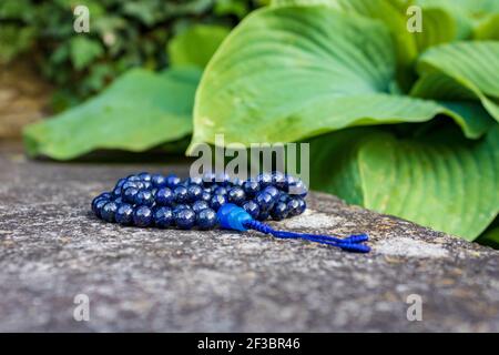 Blaue buddhistische Gebetsperlen (Lapislazuli) auf Stein mit grünen Blättern im Hintergrund. Yoga und Meditation Zubehör. Stockfoto