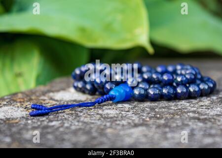 Blaue mala Perlen (Lapislazuli) auf Stein mit grünen Blättern im Hintergrund. Yoga und Japa Meditation Zubehör. Stockfoto