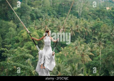 Gebräunte schöne Frau in einem langen weißen Kleid mit einem Zug, Reiten auf einer Schaukel. Im Hintergrund ein Regenwald und Palmen. Speicherplatz kopieren. Stockfoto