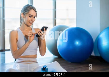 Fröhliche schöne Frau in weißer Sportbekleidung nach dem Training sitzt Auf Yoga-Matte und nutzt Telefon Stockfoto