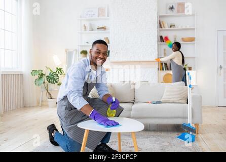 Lächelnder schwarzer Kerl Wischtisch im Wohnzimmer, während seine Freundin Staub auf Hintergrund, leeren Raum Stockfoto