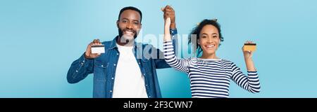 Fröhliches afroamerikanisches Paar, das Kreditkarten zeigt und dabei die Hände auf blauem Banner hält Stockfoto