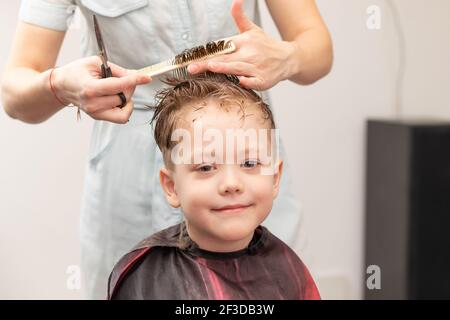 Fürsorgliche weibliche Hände einer Mutter mit Kamm und Schere machen einen modischen Haarschnitt für ihren Sohn zu Hause während der zweiten Sperre. Selektiver Fokus Stockfoto