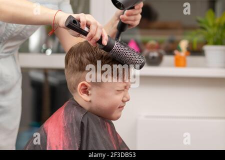 Eine Mutter mit einem Haartrockner in der Hand in einem hellblauen Kleid macht ihr Sohn während der zweiten Quarantäneperiode zu Hause die Haare. Selektiver Fokus Stockfoto