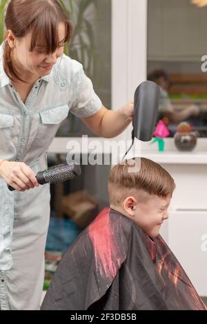 Eine Mutter mit einem Haartrockner in der Hand in einem hellblauen Kleid macht ihr Sohn während der zweiten Quarantäneperiode zu Hause die Haare. Selektiver Fokus Stockfoto