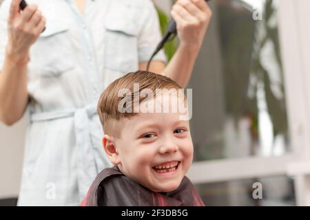 Eine Mutter mit einem Haartrockner in der Hand in einem hellblauen Kleid macht ihr Sohn während der zweiten Quarantäneperiode zu Hause die Haare. Selektiver Fokus Stockfoto