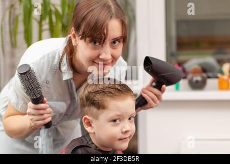 Eine Mutter mit einem Haartrockner in der Hand in einem hellblauen Kleid macht ihr Sohn während der zweiten Quarantäneperiode zu Hause die Haare. Selektiver Fokus Stockfoto