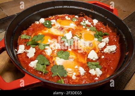 Ein köstlicher Topf mit Shakshuka mit Avocado und Harissa Eiern Kochen in einem großen roten Topf Stockfoto