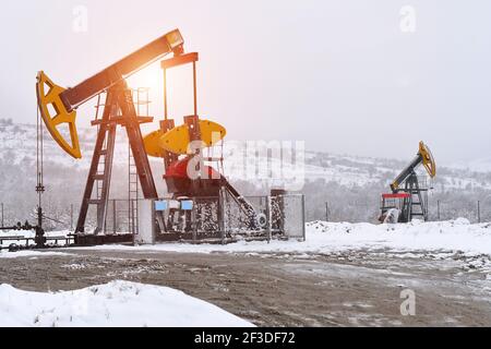 Ölpumpe. Anlagen der Öl- und Gasindustrie. Ölfeld-Pumpenheber und Ölraffinerie im Winter mit Schnee, Bergen, Wald und Sonnenuntergang im Hintergrund Stockfoto