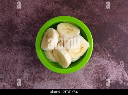 Draufsicht auf eine kleine grüne Schale, gefüllt mit frischen Bananenscheiben auf einer dunkelrot-melierten Tischplatte. Stockfoto