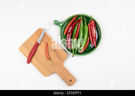 Grüne und rote Chili-Paprika auf grünem Teller, rote Paprika auf Holzbrett mit Messer, weißer Hintergrund, Schneidebrett, flaches Lay. Würze Stockfoto