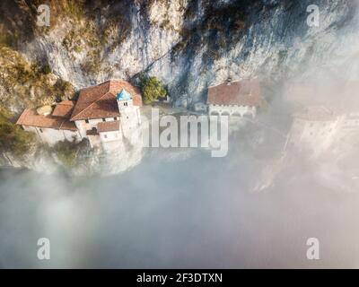 Luftaufnahme von historischen Häusern mit rotem Dach unter vertikaler Felswand. Sonne scheint durch spärlichen Nebel. Stockfoto