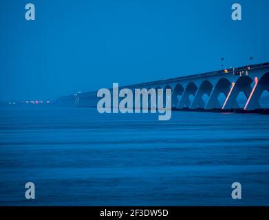 Blue Hour Blick auf lange Betonbrücke, die über die Wasseroberfläche führt. Szene bei schwachem Licht. Pfeiler, die Bögen unterstützen, bilden geometrische Formen. Stockfoto