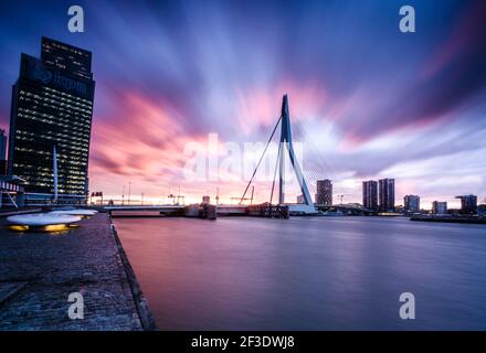 Moderne Designbrücke und mehrere hohe Gebäude am Flussufer. Sonnenuntergang Farbe Himmel mit Wolken. Lange Belichtungszeit. Stockfoto