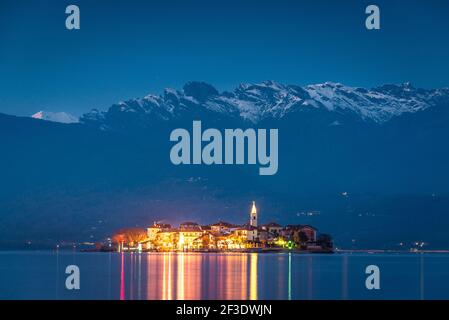 Abendansicht auf der Insel mit beleuchteten Gebäuden. Bunte Lichter, die sich auf der Wasseroberfläche des Sees spiegeln. Schneebedeckte Berge im Hintergrund. Stockfoto