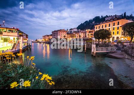 Abend am See. Die Fähre wartet auf Passagiere gegenüber von Hotels und dem von orangefarbenem Licht beleuchteten Haus. Touristenziel nach Sonnenuntergang. Stockfoto