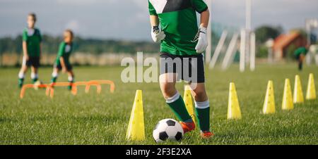 Kid Fußballspieler Dribbbling durch Kegel. Junge in Fußball Uniform üben mit Ball. Kind tritt Ball auf Gras. Junger Athlet verbessert Fußball dri Stockfoto