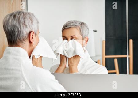 Ältere Frau wischte Gesicht mit Handtuch, während sie in der Vorderseite stand Eines Spiegels Stockfoto