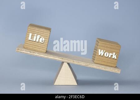 Die Holzskala mit Blöcken, die die gebrochene Balance zwischen zeigen „Leben“ und „Arbeit“ mit dem schwereren „Arbeit“-Block auf dem Blauer Hintergrund Stockfoto