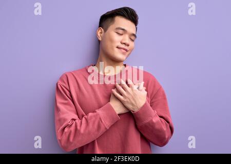 Chinesischer junger Mann, der Dankbarkeit zum Ausdruck bringt, die Hände auf der Brust hält und die Augen isoliert auf violettem Hintergrund geschlossen hält, Porträt eines Mannes, der Generos fühlt Stockfoto