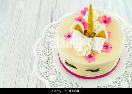 Einhorn-Kuchen auf Holzhintergrund und Spitze. Niedliche Einhorn geschichteten hausgemachten Kuchen mit Schleife, Rosenblumen, goldenen Mais und Ohren verziert. Speicherplatz kopieren. Stockfoto