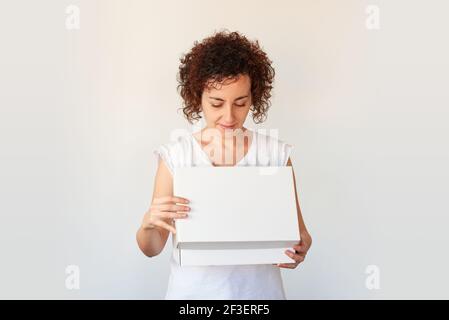 Eine junge Frau mit lockigen Haaren öffnet eine weiße Schuhbox, die sie in einem Online-Shop bestellt hat. Das Unternehmen liefert Pakete schnell zu Ihnen nach Hause. Anniversa Stockfoto