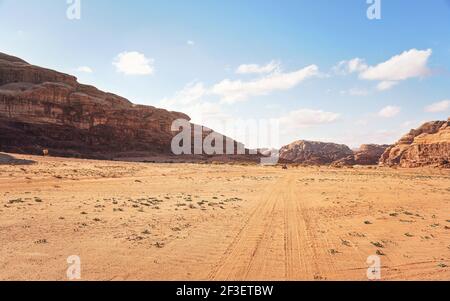 Felsige Massive auf roter Sandwüste, heller wolkiger Himmel im Hintergrund, kleines 4WD Fahrzeug in der Ferne - typische Landschaft im Wadi Rum, Jordanien Stockfoto