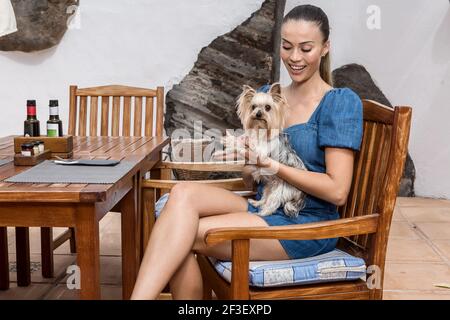 Glückliche Frau lächelt und hält niedlichen Yorkshire Terrier während des Sitzens In der Nähe des Tisches im Außenrestaurant des hundefreundlichen Hotels Stockfoto