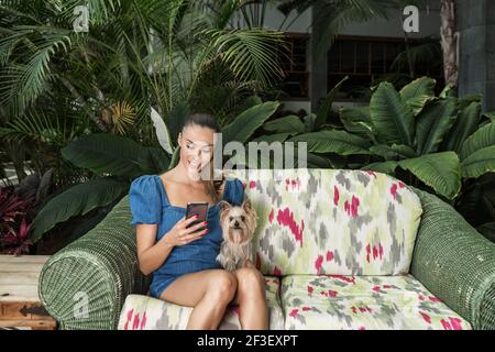 Schöne Dame mit niedlichen Yorkshire Terrier lächeln und browsen Smartphone Beim Sitzen auf weicher Couch in der Nähe von grünen Pflanzen im Hund Freundliches Hotel Stockfoto