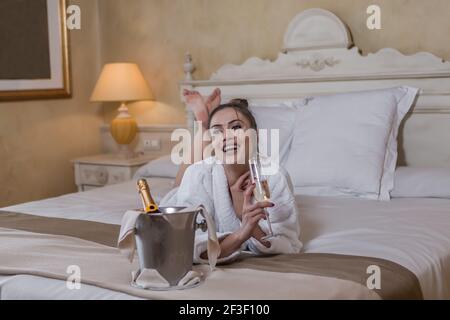 Aufgeregt attraktive Frau in weißem Bademantel lachend und wegschauen Beim Champagner trinken und auf dem Bett in der Nähe von Eimer mit liegen Kalte Flasche in Luxus Stockfoto