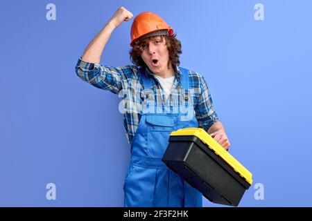 Junge männliche Ingenieur in blauen Overalls klopft auf orange Helm, sicherzustellen, Mitarbeiter arbeiten mit gutem Schutz, lila Studio Hintergrund, Porträt von b Stockfoto