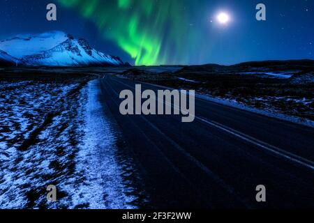 Die isländische Ringstraße Hringvegur (Route 1) führt in Richtung der schneebedeckten Vestrahorn-Berge, unterhalb der Nordlichter und des Mondes. Stockfoto