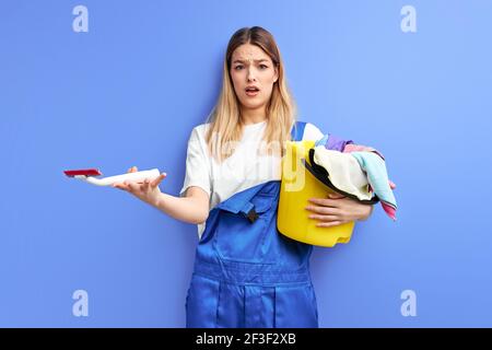 Frau hält Eimer mit Gruppe von Reinigungsmitteln. Junge Dame in Overalls stehen in Schock durch Schmutz, Haushaltsgeräte, Frühjahrsputz, Aufräumen Stockfoto