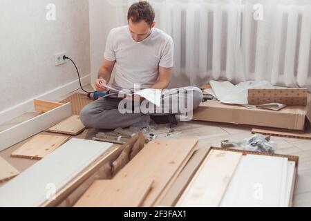 Verwirrter Mann, der zu Hause neue Holzmöbel zusammengebaut hat. Mann liest Anweisungen und missversteht, was als nächstes zu tun. Stockfoto