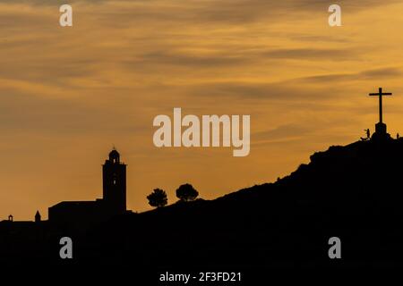 Silhouette von Menschen und Hund auf der Oberseite eines Hügel mit einem großen Kreuz mit einer Kirche und Bäumen Im Hintergrund während der letzten Lichter der Dämmerung Stockfoto