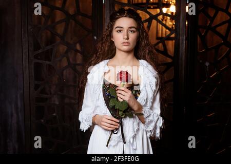 Eine junge schöne Brünette mit langen lockeren Haaren in einem Weißer Peignoir in der Art und Weise des 19th. Jahrhunderts hält Eine scharlachrote Rose in ihren Händen - ein Cosplay für Das Broadway Musical das Phantom der Oper Stockfoto