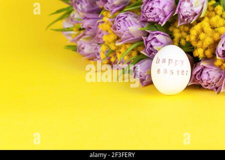 Ein weißes Hühnereier mit Worten Frohe Ostern geschrieben mit Hilfe von Briefmarken und rosa Metallic-Farbe, Bouquet von Tulpen und Mimosen auf gelbem Hintergrund, Stockfoto