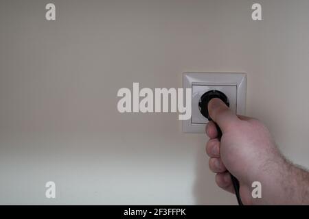 Eine männliche Hand zieht an einem schwarzen Netzkabel, das eingesteckt ist In eine Steckdose Stockfoto