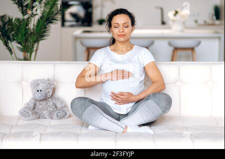 Erstaunliche gemischte Rasse Erwachsene schwangere Frau trägt weißes T-Shirt und Leggings entspannt im Wohnzimmer auf dem Sofa zu Hause, meditiert in der Lotus-Position, sanft umarmt ihren Bauch, denken Sie an Baby, Augen geschlossen Stockfoto