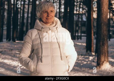 Porträt einer glücklichen Frau mittleren Alters im Winter in einem schneebedeckten Stadtpark bei Sonnenuntergang. Winterurlaub und Reisekonzept. Stockfoto