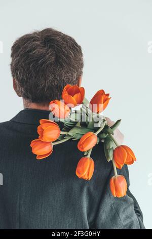 Ein eleganter Mann mittleren Alters trägt einen Strauß oranger Tulpen auf seiner Schulter. Überraschung für Ihre geliebte Frau. Romantisches Kurzurlaub-Konzept. Rückansicht. Stockfoto