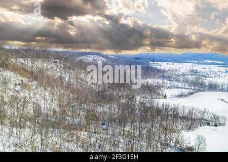 Ein dynamischer Himmel mit Sonnenstrahlen über einer verschneiten Winterlandschaft in Jackson County, IN. Schneeverwehung tritt in zwei Bereichen am fernen Horizont auf. Stockfoto