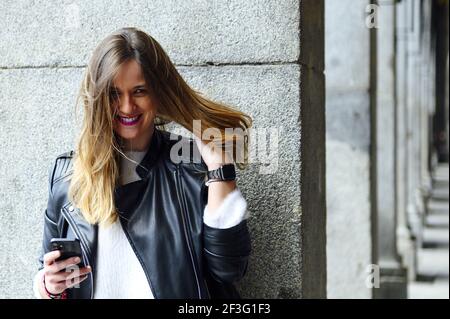 Ein Porträt einer verrückten jungen Spanierin, die sie fixiert Haare lehnen sich an eine Steinsäule, die ihr Telefon hält Stockfoto