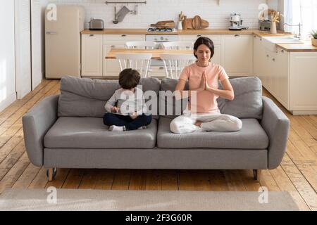 Mutter macht Yoga-Übung, sitzen auf der Couch zu Hause, Kind mit Gadget, spielen ein Spiel auf Tablet. Stockfoto