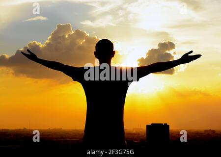 Silhouette eines Mannes, der seine Arme auf dem Hintergrund des Dämmerhimmels hebt - glückliche, entspannte & erfolgreiche Konzepte Stockfoto