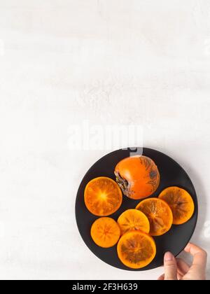 Reife orange saftige Kaki Scheiben und ganze Frucht auf schwarzem Teller auf weißem strukturiertem Hintergrund. Draufsicht. Kopierbereich. Stockfoto