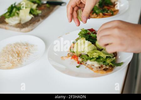 Hände legen Salatscheiben auf hausgemachte Pizza auf Teller in der modernen Küche. Scheibe leckere Pizza mit Pilzen, Tomaten, Rucola und Käse auf dem Teller. Stockfoto